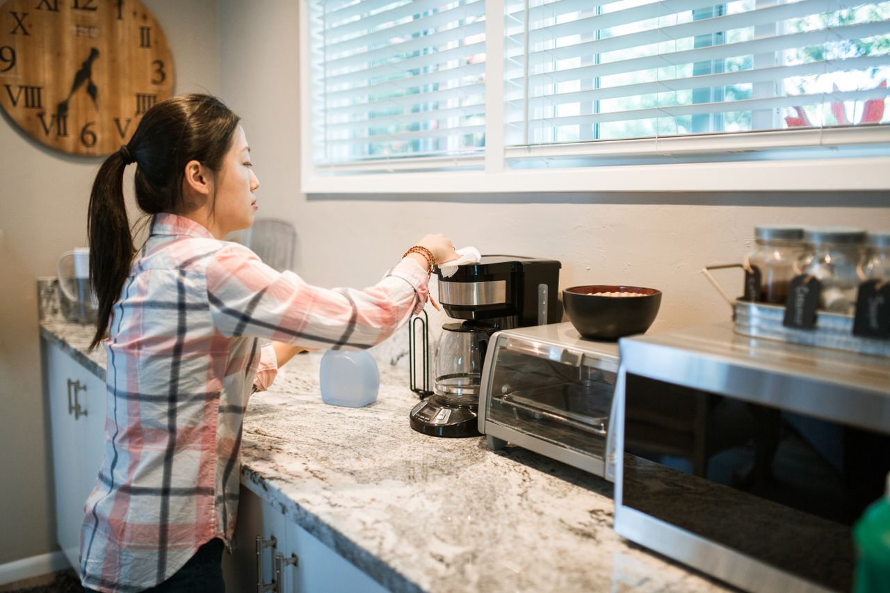 services-01 Woman cleaning a coffee maker in a bright, modern kitchen with appliances.