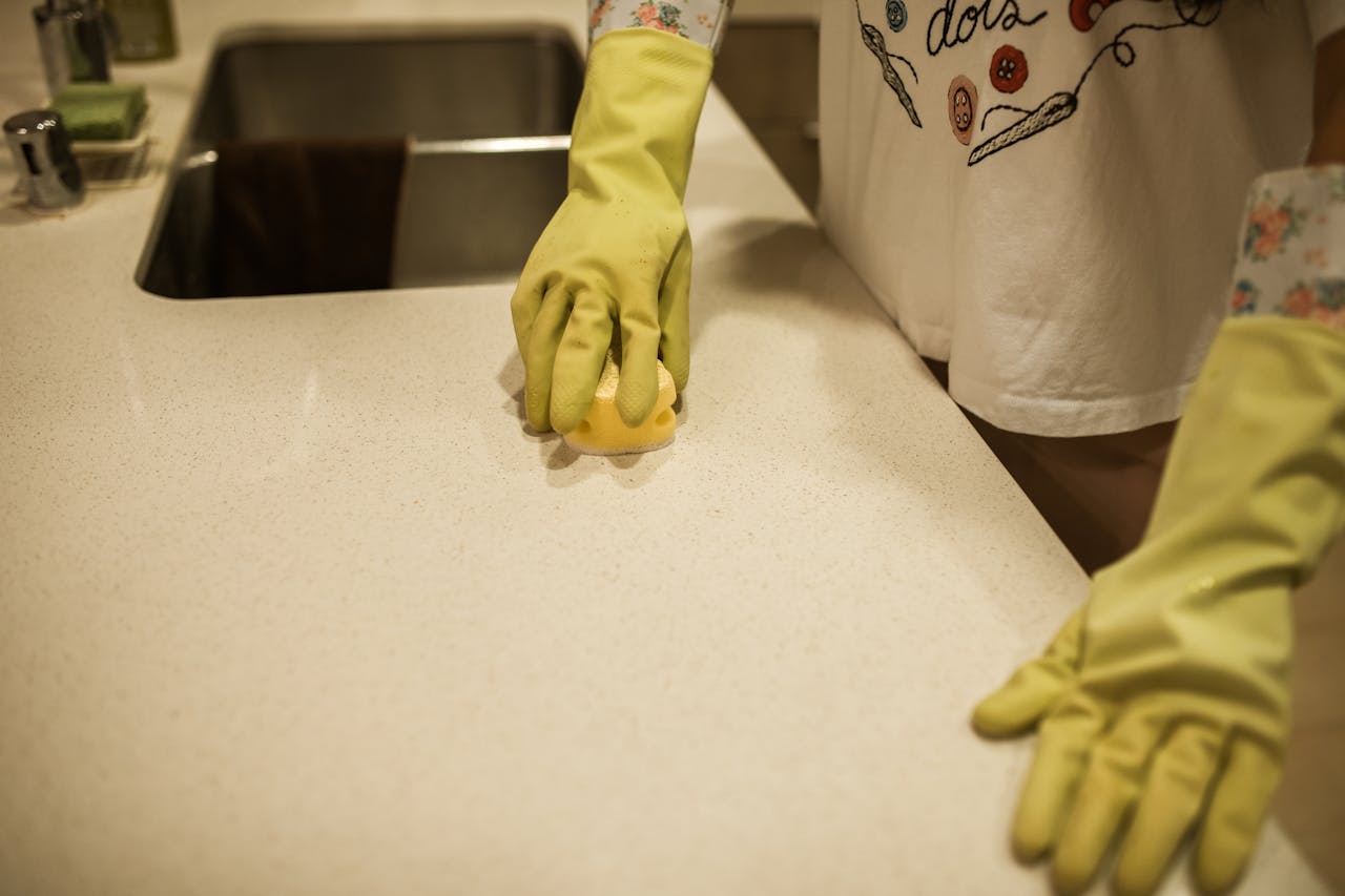 hero-img-01 Woman using yellow rubber gloves to clean a kitchen counter with a sponge.