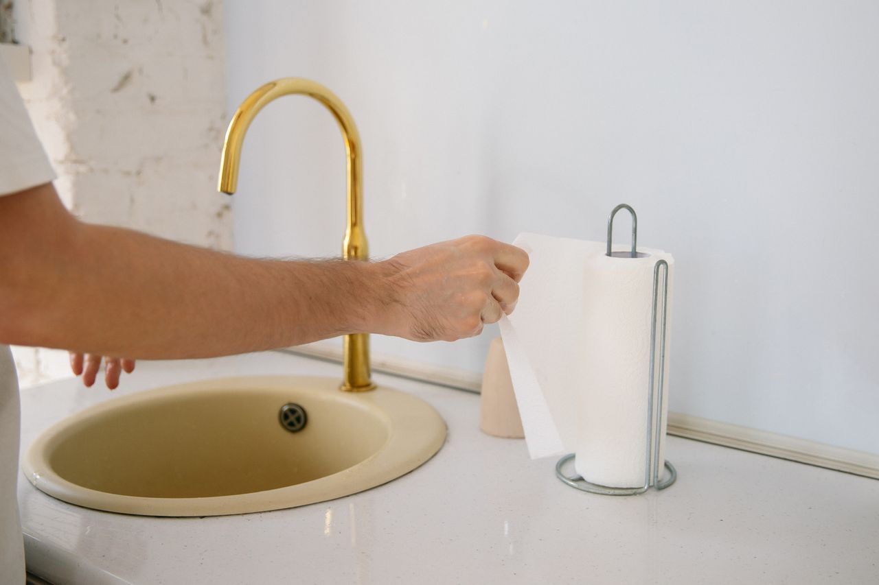 hero-img-02 A bathroom sink with a golden faucet and hand reaching for a paper towel in a clean setting.